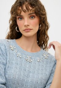 Maglione lavorato a maglia azzurro chiaro con un motivo testurizzato e dettagli floreali in perle intorno al collo. Maniche corte e vestibilità rilassata.