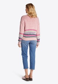Rosa Strickpullover mit mehrfarbigen horizontalen Streifen an der Taille, kombiniert mit hellblauen, knöchellangen Jeans und rosa, Nieten-besetzten Clogs.