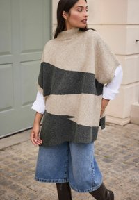 Beige and dark gray patterned knit poncho with a high collar, paired with light blue wide-leg denim shorts and brown knee-high boots.