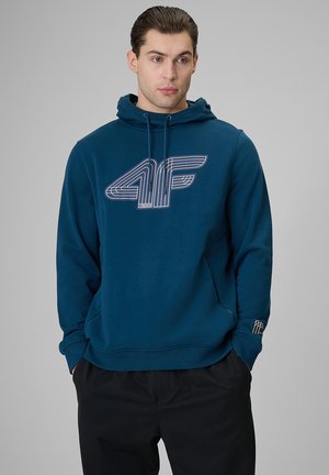 Mężczyzna w niebieskiej bluzie z kapturem z dużym stylizowanym logo "4F" na piersi oraz czarnych spodniach, ręce w kieszeniach, neutralne tło.
