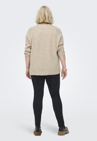 Beige gerippter Strickpullover mit lockerem Schnitt, überschnittenen Schultern und abgerundetem Saum, kombiniert mit schwarzen Figur betonten Hosen und schwarzen Schuhen.