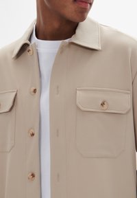 Camisa de botones beige hecha de tela suave, con un cuello clásico, dos bolsillos en el pecho con cierres de botón, y una capa interna blanca.