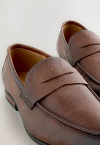 Braune Lederloafers mit strukturiertem Finish, ausgestattet mit einem Riemen über der Oberseite und einer flachen schwarzen Sohle mit leichtem Absatz.