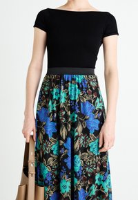 Svart ribbad topp med kort ärm i kombination med en blommig kjol med blå och gröna mönster på svart bakgrund. Modellen håller en brun väska.