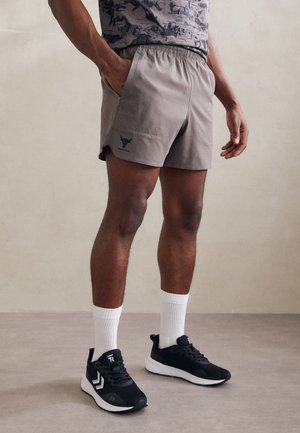 Mann trägt graue Sportshorts mit einem Logo, schwarze Sneakers, weiße Kniestrümpfe und ein gemustertes Hemd, steht mit einer Hand in der Tasche.
