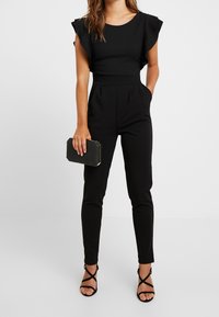 Svart jumpsuit med korta volangärmar, figurnära midja och avsmalnande ben. I kombination med en svart rektangulär clutch och sandaler med remmar.