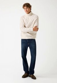 Cremefarvet kabelstrikket rullekravesweater, kombineret med mørkeblå jeans med lige ben. Sweateren har teksturerede mønstre og ribbede ærmekanter.