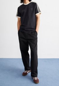 Muž v černém tričku Adidas s bílými pruhy, černých kalhotách a hnědých teniskách stojící na modrém koberci před bílou stěnou.