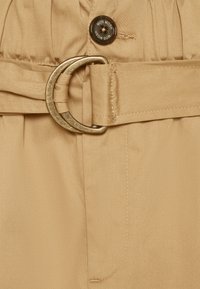 Tessuto beige con una superficie liscia, caratterizzato da una grande fibbia metallica circolare e un bottone con il logo "MOS MOSH". Presenti dettagli cuciti.