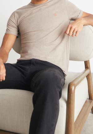 T-shirt beige ajusté avec un petit logo orange sur la poitrine, associé à un jean noir. Le décor comprend un fauteuil clair texturé.