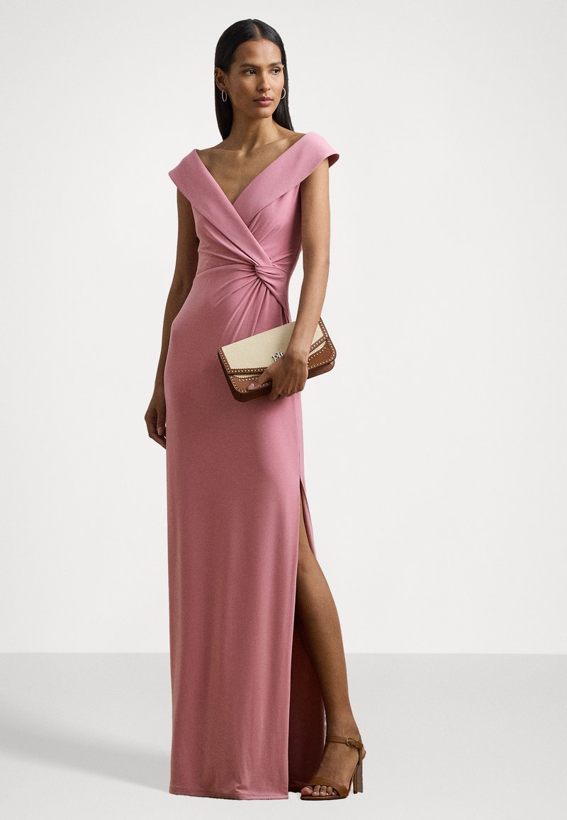 Langes pinkes Kleid mit schulterfreiem Design, gedrehten Frontdetails und seitlichem Schlitz. Gestylt mit einer beigen Clutch und braunen Riemchen-Heels.
