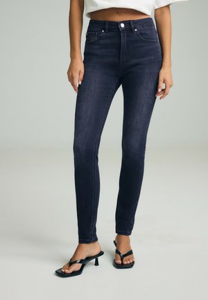 Kvinde iført mørkeblå skinny jeans med høj talje, hvid cropped top og sorte hæle sandaler på en ensfarvet baggrund.