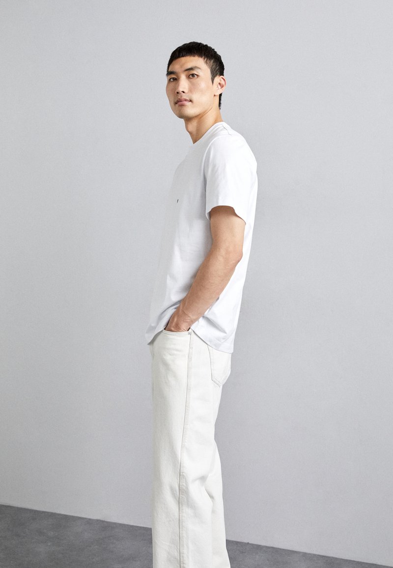 White cotton t-shirt with short sleeves, ribbed crew neckline, and a small logo. Paired with light-colored, wide-leg jeans.