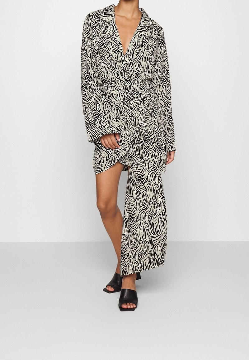 Langes, zebra-musterndes Kleid in Creme und Schwarz mit tiefem V-Ausschnitt, lockeren Ärmeln und hohem seitlichem Schlitz, kombiniert mit schwarzen Slipper-Sandalen.