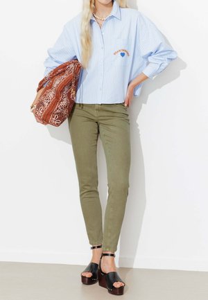 Femme portant une chemise à rayures bleu clair et un pantalon vert olive tient un grand sac marron à motifs, debout sur fond blanc.