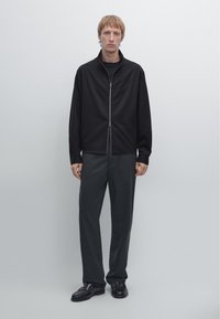 Svart zip-up jacka med hög krage, kombinerad med mörkgrå byxor och svarta skor. Enkelt, slätt tyg och skräddarsydd passform.