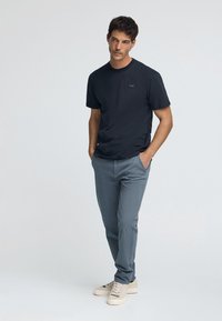 Dunkelblauses T-Shirt mit kurzen Ärmeln und kleinem Patch-Logo, kombiniert mit hellgrauen Hosen und beigen Sneakern. Glatte Stofftextur über das gesamte Kleidungsstück.