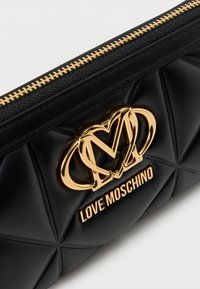 Μαύρο τσαντάκι από συνθετικό δερματίνη με καπιτονέ σχέδιο και εξαρτήματα σε χρυσό τόνο, με εμφανές λογότυπο και φερμουάρ στην κορυφή.