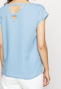 Ljusblå denimtopp med rund halsringning, uppvikta ärmar och en nyckelhålskärning på baksidan. Mjuk textur och avslappnad passform.