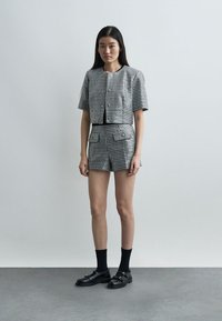 Jeune femme debout portant une veste à manches courtes en pied-de-poule noir et blanc assortie à un short taille haute, avec des chaussures et des chaussettes noires.