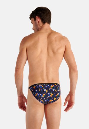 Gespiervde man van achteren gezien, gekleed in zwarte zwemslips met een kleurrijk abstract bloemmotief, staand tegen een effen lichte achtergrond.