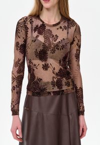 Vrouw die een transparante top met lange mouwen en donkere bloemmotieven draagt over een bruine beha en een donkerbruine lederen rok.