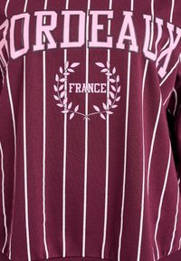 Bordeaux pulover v burgundski barvi z belimi vertikalnimi črtami. Ima napis "BORDEAUX" in "FRANCE" v roza barvi, obdan z laurelnim motivom.