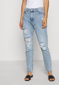 Jasnoniebieskie, wysokie spodnie dżinsowe z przetarciami, noszone z czarnymi tkanymi sandałami na płaskiej podeszwie i białą bluzką, skróconą do wysokości połowy torsu, na jednolitym tle.