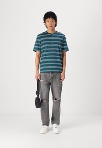 Camiseta de manga corta a rayas en tonos azul verdoso, jeans grises rasgados y zapatillas blancas. Lleva un pequeño bolso negro cruzado. Diseño simple, ajuste casual.