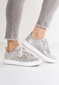 Stopy w jasnoszarych, błyszczących sneakersach z satynowymi sznurowadłami i perłowymi ozdobami na obcasie, w połączeniu z poszarpanymi, szarymi dżinsami.