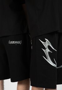 Black t-shirts and shorts, featuring silver accents. Shorts have a side design and branding on pockets, highlighting textured fabric.