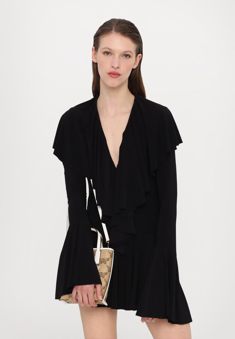 Zwarte wikkeljurk met gerimpelde mouwen en een diepe V-hals. Model houdt een gestreepte handtas vast met een witte strap. Geverweven stof.