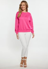 Sudadera rosa con mangas texturizadas adornadas con pequeños abalorios decorativos, combinada con pantalones ajustados blancos y sandalias de tacón con tiras.