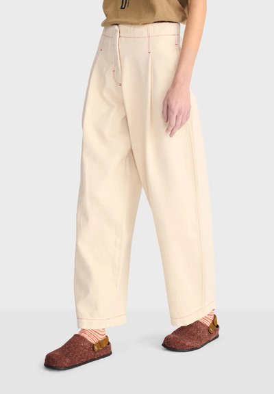 Personne portant un pantalon ample crème avec des coutures rouges, des chaussettes rayées et des chaussures marron à enfiler, debout sur une surface gris clair.