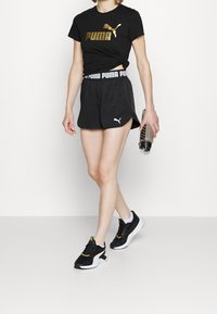 Žena kráčející ve černých sportovních šortkách Puma, s vázaným černým tričkem Puma, černobílých teniskách a držící v pravé ruce láhev s vodou.