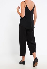 Black sleeveless top with a V-back, paired with loose-fitting black cropped trousers and flat black shoes. Smooth fabric, minimal design.