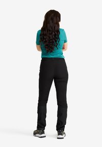 Kvinna står med armarna korsade, iförd en teal t-shirt med kort ärm, svarta byxor och vandringsskor, vänd bort mot en vit bakgrund.