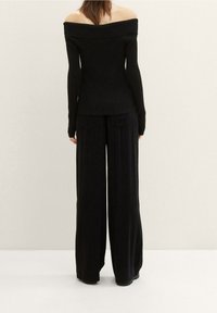 Femme debout, dos tourné vers le spectateur, portant un haut noir à manches longues décolleté sur les épaules et un pantalon large noir, sur fond uni.