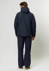 Mann in einer marineblauen Kapuzenjacke und passenden Hosen, der mit dem Rücken zur Kamera auf einem schlichten hellgrauen Hintergrund steht.