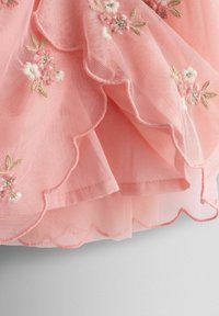 Rosa bestickter Tüllrock mit gewelltem Saum, verziert mit zarten Blumenmustern in weißem und grünem Garn auf einem weichen Hintergrund.