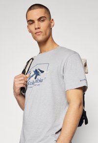 Camiseta de algodón gris con un gráfico de montaña azul y el texto "Columbia". El modelo sostiene una mochila de tira negra. Cuello redondo simple y mangas cortas.
