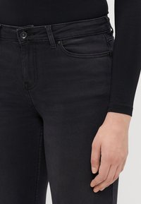 Juodos džinsinės kelnės su liesu siluetu, turinčios standartinį penkių kišenių dizainą, mygtuko užsegimą ir subtilias siuvimo detales.