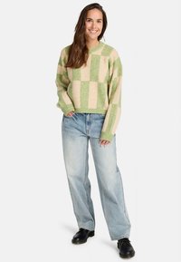 Šypsena moteris, vilkinti žalią ir smėlio spalvos languotą megztinį, laisvas šviesiai mėlynas džinsus ir juodus batus, stovi su viena ranka kišenėje.