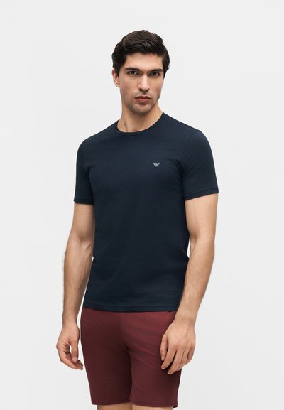 Άνδρας που φοράει εφαρμοστό κοντομάνικο μπλε μπλουζάκι και μπορντό σορτς, στέκεται μπροστά σε ένα απλό ανοιχτό φόντο.