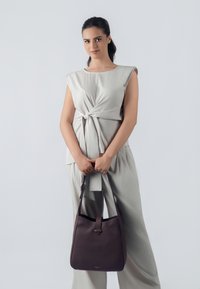 Borsa a tracolla in pelle marrone con una texture liscia e manico corto. Indossata da una modella in un outfit grigio chiaro con un design anteriore attorcigliato.