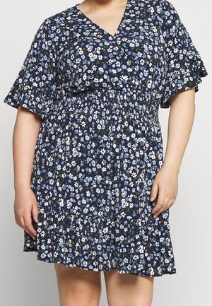 Femme portant une robe bleu marine avec un petit imprimé floral blanc et bleu clair, des manches courtes volantées, un col en V, et une taille et un ourlet froncés.