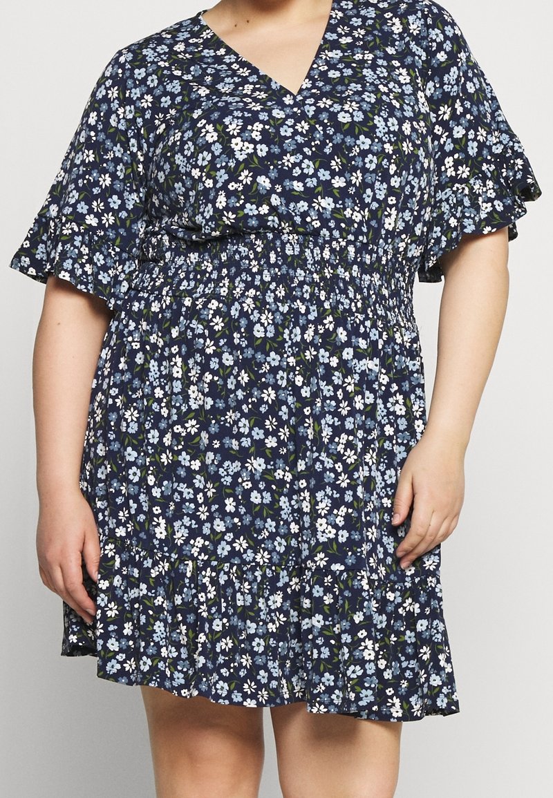 Femme portant une robe bleu marine avec un petit imprimé floral blanc et bleu clair, des manches courtes volantées, un col en V, et une taille et un ourlet froncés.