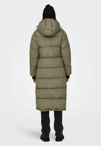 Olivgrüner Puffercoat mit Kapuze, mit einem langen, gesteppten Design und verstellbaren Kordelzügen. Getragen mit dunkelblauen Jeans und schwarzen Stiefeln.