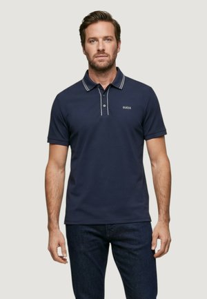 Homme portant un polo bleu marine à manches courtes avec un liseré blanc sur le col et le logo "GUESS" sur le côté gauche de la poitrine.