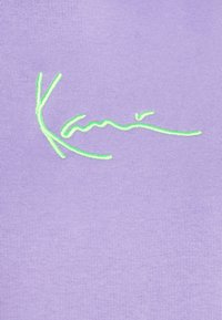 Texto "Kanié" bordado en verde sobre tela acanalada de color púrpura claro, resaltando el logotipo de la marca en el material de la ropa.